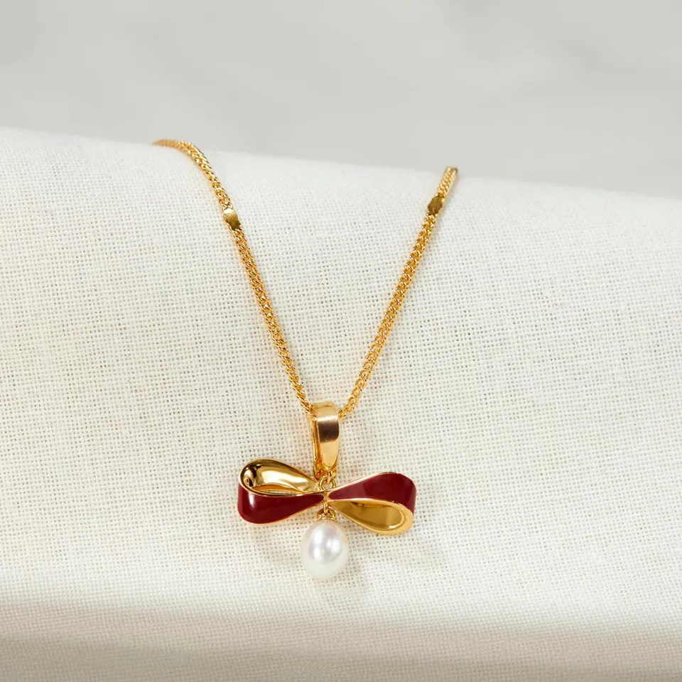 18K gold vermeil burgundy enamel bow pendant with a natural pearl drop, resting on cream linen